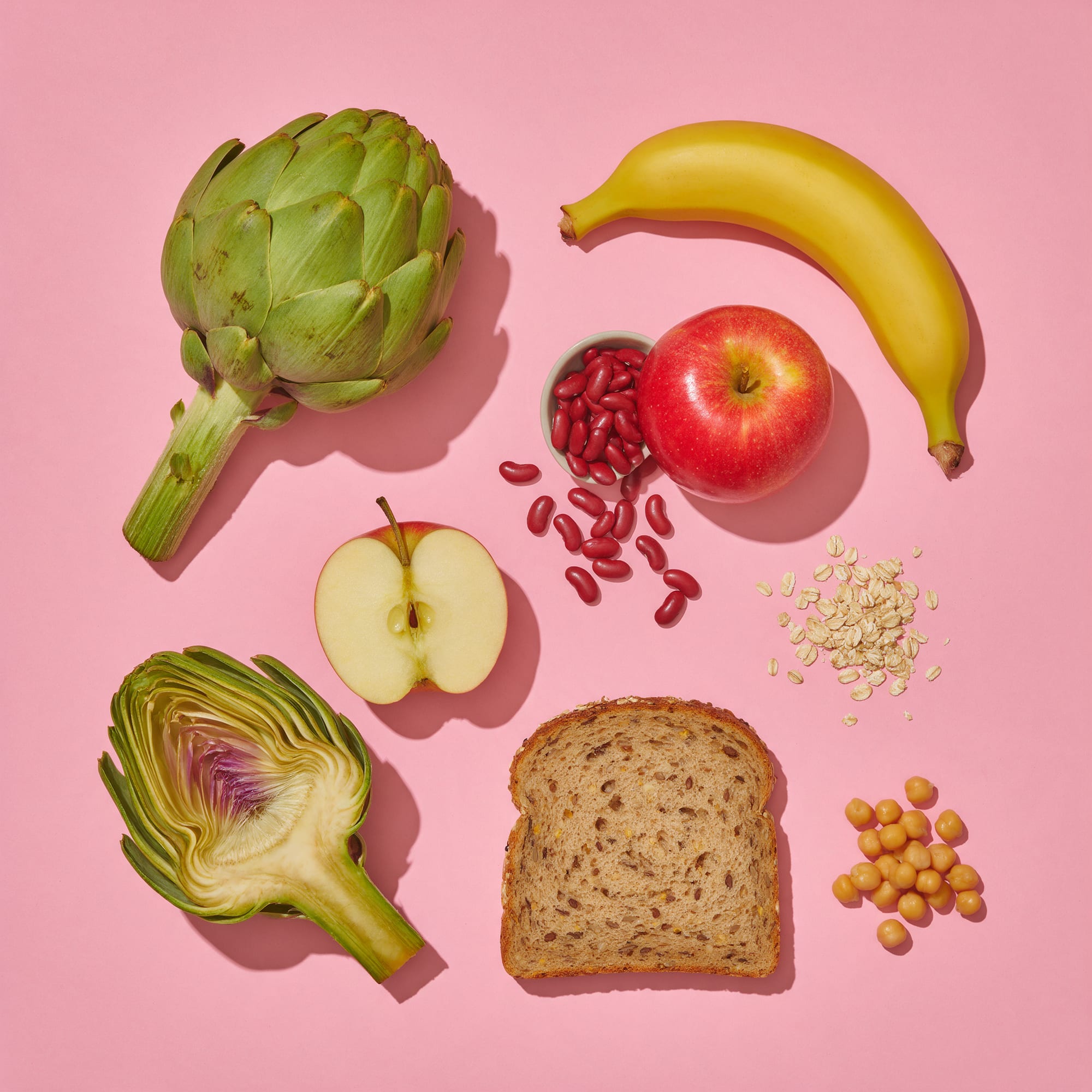 Alcachofas, plátano, manzana, alubias, avena, pan integral y garbanzos sobre fondo rosa
